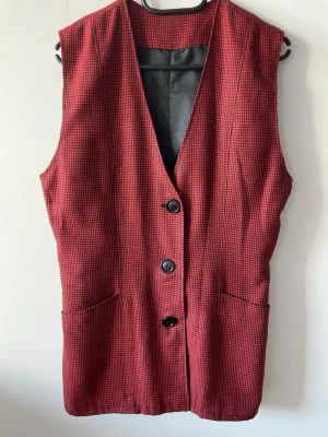 Rutig röd väst med knappar - Röd väst med svart-rutigt mönster och tre svarta knappar framtill. Västen har två fickor och en djup v-ringning. 