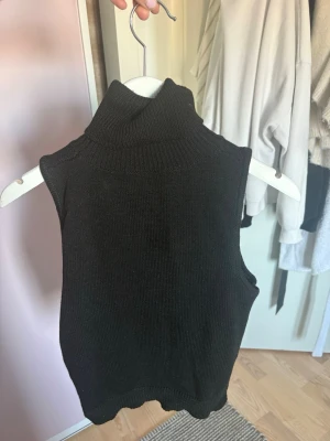 Svart stickad väst med polokrage 🖤 - Säljer en svart stickad väst med hög polokrage. Västen har en enkel och clean design utan ärmar och är perfekt att styla över en skjorta eller t-shirt. Ribbad kant nedtill och runt kragen ger en snygg finish.