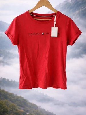 Röd t-shirt från Tommy Hilfiger - Snygg korall t-shirt från Tommy Hilfiger med klassisk logga tryckt på bröstet. T-shirten har rund halsringning och korta ärmar. Tillverkad i mjuk bomull som känns skön mot huden. Perfekt för en avslappnad och stilren look.