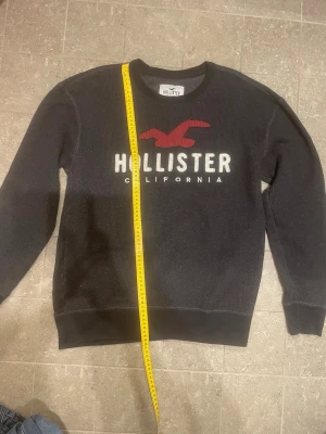 Svart Hollister sweatshirt med logga - Svart sweatshirt från Hollister med stor vit och röd logga på bröstet samt texten 'California'. Tröjan har rund halsringning, ribbade muddar och är tillverkad i mjukt material. Perfekt för en avslappnad och trendig stil.sthlm m