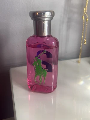 Ralph Lauren Big Pony Pink Parfym - En parfymflaska från Ralph Lauren Big Pony Collection med rosa glas och silverfärgad metallkork. Flaskan har en grön polospelare och en stor lila siffra 2 på framsidan. 