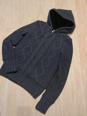Mörkgrå stickad cardigan med dragkedja - Mysig mörkgrå stickad hoodie med dragkedja framtill och huva med dragsko. Tröjan har ett diskret kabelstickat mönster på framsidan och breda ribbade muddar vid ärmslut och nederkant. Perfekt för kalla dagar och enkel att matcha. Storlek S sitter mindre som xs
