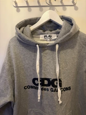 Grå hoodie Comme des Garçons Play - Snygg grå hoodie från Comme des Garçons Play med svart CDG-tryck på bröstet. Klassisk känguruficka framtill, vita dragsnören i huvan och mjukt material. Perfekt för en chill och trendig look! Nypris 2000  kr mitt pris 450 kr 