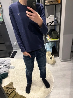 Marinblå långärmad topp från Gant - En stilren marinblå långärmad topp från Gant med rund halsringning och diskret logga broderad på bröstet. Tillverkad i mjuk bomull som känns skön mot huden. Perfekt att matcha med jeans eller kjol för en clean look.