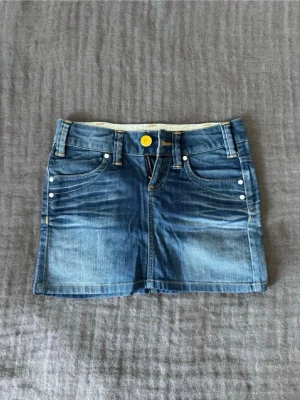 Blå jeanskjol med gul knapp - Snygg blå lågmidjad jeanskjol med jätte skön passform. Kjolen har en kort modell och har perfekt längd. Tyvärr är den för liten för mig nu så därför säljer jag. Kjolen är i jätte bra skick och super snygg till våren och sommar! Fråga om mått!