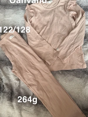 Rosa ribbad pyjamas med volangdetaljer - Mysig tvådelad pyjamas i ljusrosa ribbat tyg. Tröjan har långa ärmar och söta volangdetaljer vid axlarna. Byxorna är raka och mjuka, perfekta för att chilla hemma. Storlek 122/128, passar barn eller ungdomar.🌸 Ny utan lapp. Storlek 122/128