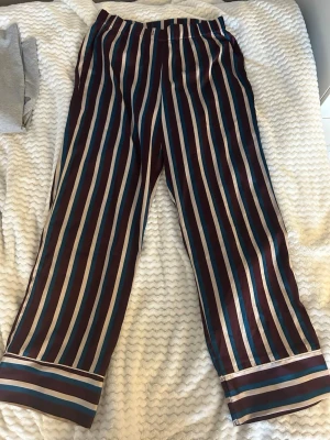 Randiga pyjamasbyxor från Gina Tricot - Säljer ett par pyjamasbyxor från Gina Tricot med breda ränder i vinrött, blått och vitt. Byxorna har resår i midjan och är gjorda i ett mjukt, glansigt material som känns lyxigt mot huden. Perfekta för mysiga hemmakvällar.