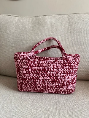 Rosa och vinröd virkad handväska - Unik handväska i virkad stil med två handtag, gjord av tjockt garn i rosa och vinrött. Väskan har en rektangulär form och ger en trendig, handgjord känsla. Perfekt accessoar för dig som vill sticka ut med färg och textur.