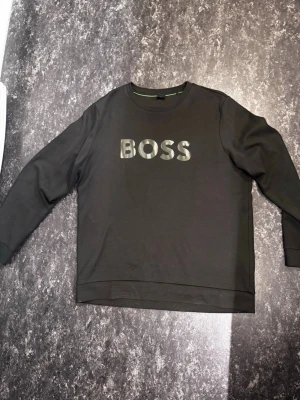 Hugo boss - Snygg svart sweatshirt från BOSS med stor logga i blankt tryck på bröstet. Tröjan har rund halsringning, långa ärmar och ribbade muddar. Materialet är mjukt och bekvämt, perfekt för en avslappnad och trendig stil.