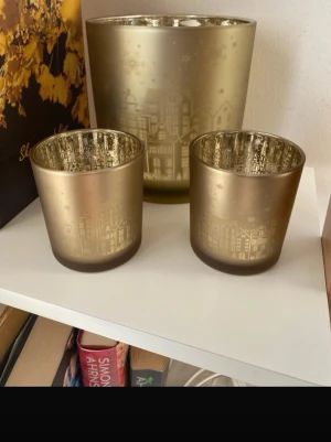 Guldiga ljuslyktor med stadsmotiv nya  - Tre guldiga ljuslyktor i olika storlekar med ett snyggt stadsmotiv runt om. Insidan är spegelblank vilket ger ett mysigt sken när ljusen är tända. Perfekta som dekoration på hyllan eller bordet. Materialet är glas och färgen är metallic guld.