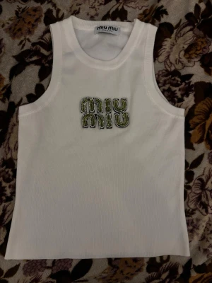 Vit ärmlös topp från Miu Miu - Snygg vit ärmlös topp från Miu Miu med broderad logga framtill i grönt och silver. Tillverkad i ett ribbat material som ger en trendig och fräsch look. Perfekt att styla med jeans eller kjol för en cool vibe.