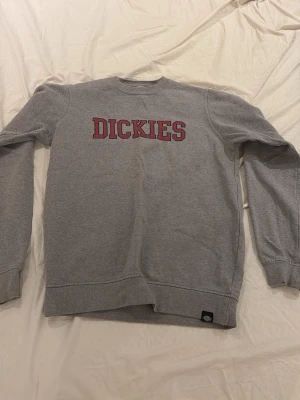 Grå sweatshirt från Dickies - Säljer en grå sweatshirt från Dickies med röd logga broderad på bröstet. Tröjan har rund halsringning, ribbade muddar vid ärmslut och nederkant samt mjuk insida i fleece. Perfekt för en avslappnad och stilren look.