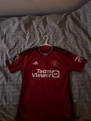 Manchester United matchtröja Garnacho - Säljer en röd Manchester United matchtröja från Adidas med Garnacho och nummer 17 på ryggen. Tröjan har svarta detaljer, klubbmärke på bröstet, Premier League-märke på ärmen och TeamViewer som sponsor framtill. Perfekt för fotbollsfans! 