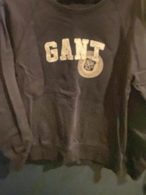 Svart sweatshirt från GANT - Klassisk svart sweatshirt från GANT med stor vit logga och emblem på bröstet. Tröjan har rund halsringning och långa ärmar. Perfekt för en avslappnad och sportig stil. Tillverkad i mjukt material för extra komfort.