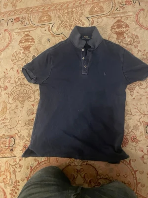 Marinblå pikétröja från Polo Ralph Lauren - Klassisk marinblå pikétröja från Polo Ralph Lauren med korta ärmar, krage och knappar framtill. Tröjan har den ikoniska broderade loggan på bröstet och är tillverkad i mjuk bomull. Perfekt för en avslappnad men stilren look.