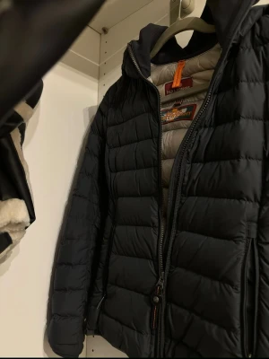 Svart dunjacka från Parajumpers - Snygg svart dunjacka från Parajumpers med quiltad design och dragkedja framtill. Jackan har huva och detaljer i orange vid halsen. Perfekt för kalla vinterdagar och riktigt skön att bära.