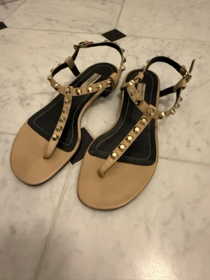 Beige sandaler med nitar från Balenciaga - Snygga beige sandaler från Bullboxer med T-rem och guldfärgade nitar. Sandalerna har justerbart spänne runt ankeln och svarta detaljer på innersulan. Perfekta för en trendig och avslappnad look.