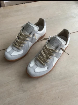 Maison Margiela gats  - Säljer ett par Maison Margiela Replica sneakers i vitt och ljusgrått med beige snörning och klassisk gummisula. Skorna har en stilren retrodesign med paneler i skinn och mocka, perfekta för dig som gillar minimalistisk och clean stil.