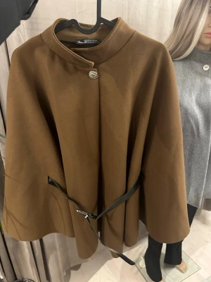 Brun cape-rock med bälte - Stilren brun cape-rock från Zara Woman med elegant knapp vid halsen och svart bälte i midjan. Rocken har en lös, svepande passform och är tillverkad i ett mjukt tyg som ger en lyxig känsla. Perfekt för dig som vill ha en trendig och unik ytterjacka.