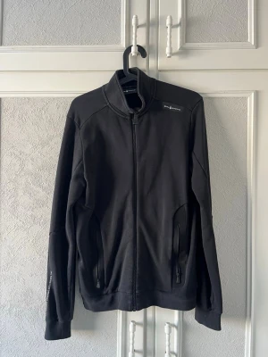 Svart kofta från sail racing - Svart kofta från sail racing med hög krage och hel dragkedja framtill. Tröjan har två fickor med dragkedja och diskret logga på bröstet. Skriv för fler bilder.