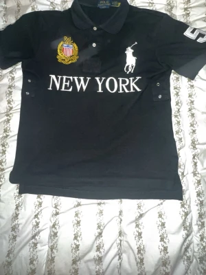 Svart Polo Ralph Lauren skjorta - Klassisk svart skjorta från Polo Ralph Lauren med vit logga, broderad patch och texten 'NEW YORK' på bröstet. Stor vit siffra 5 på ryggen och korta ärmar. Perfekt för dig som gillar sportig och stilren look.