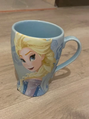 Frost Elsa mugg ljusblå - Säljer en ljusblå mugg med motiv av Elsa från Disneyfilmen Frost. Muggen har ett böjt handtag och är dekorerad med blå och vita detaljer samt ett stort tryck av Elsa i sin ikoniska klänning. Perfekt för varma drycker och för alla Frost-fans.