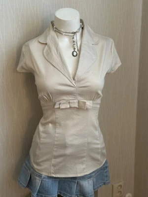 Krämvit blus med rosett från Next - cream-colored Next blouse ,a short-sleeved top featuring a notched collar and a decorative bow detail at the waist. Stretchy material tag says M but it fits more like small has som faint spots 