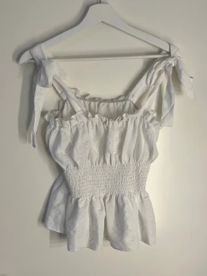 Vit linnetopp med knytband - Söt vit linnetopp med breda axelband som knyts på axlarna. Toppen har smock i midjan för en figurnära passform och volangkant upptill och nedtill. 