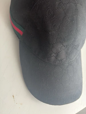 Gucci Svart keps från Gucci med GG-mönster - Säljer en svart keps från Gucci med det klassiska GG-monogrammet i tyget. Kepsen har en böjd skärm och justerbart spänne bak. På sidan finns en grön och röd rand som ger den en ikonisk look. Perfekt för dig som vill ha en stilren och lyxig accessoar.