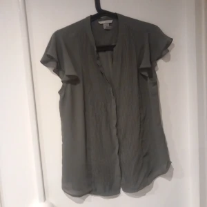 Grå plisserad blus med volangärm - Snygg grå blus med plisserad framsida och korta volangärmar. Luftigt och lätt material som ger en avslappnad men ändå chic look. Perfekt att styla med jeans eller kjol för en trendig outfit.