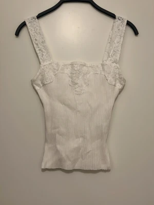 Vit spetsdetaljerad linnetopp Zara - Supersöt vit linnetopp från Zara med breda axelband och spetsdetaljer upptill. Toppen är ribbad och har en figurnära passform som ger en fräsch och somrig look. Perfekt att styla med jeans eller kjol för en trendig outfit.