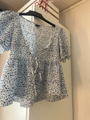 Blommig vit och blå blus med knyt - Supersöt blus med vit botten och blått blommönster. Blusen har korta puffärmar, v-ringning med volangkant och knyt framtill. Materialet är lätt och luftigt, perfekt för varma dagar. Passar till både jeans och kjol för en fräsch look.