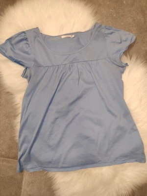 Jättefin Wera Stockholm blå topp, storlek S - Söt ljusblå topp med rund halsringning och korta puffärmar. Toppen har en lös och luftig passform med rynk under bysten för en avslappnad look. Tillverkad i mjukt bomullstyg som känns skönt mot huden.Bä Är i fint använt skick, den har en svag färgskiftning under armarna (se bild). Köpt på Åhléns, storlek S. 