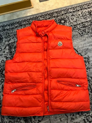 Röd dunväst från Moncler - Snygg orange väst från Moncler med hög  och två dragkedjefickor framtill. Västen är äkta och är använd en liten tid. Dragkedjan på den är sönder den går inte att dra i. Storlek 3 Medium. Vid snabb och smidig affär kan jag gå ner i priset