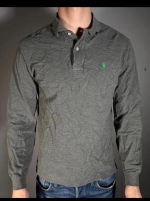 Grå långärmad skjorta från Ralph Lauren - Snygg grå långärmad skjorta från Ralph Lauren med klassisk krage och knappar framtill. Skjortan har en broderad grön logga på bröstet och är tillverkad i mjuk bomull. Perfekt för en stilren och avslappnad look.