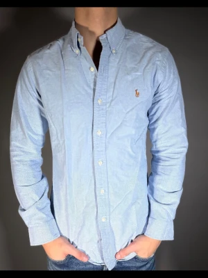Ljusblå skjorta från Ralph Lauren - Klassisk ljusblå skjorta från Ralph Lauren med button-down krage och broderad logga på bröstet. Skjortan har lång ärm, knappar framtill och är tillverkad i bomull. Perfekt för en clean och stilren look.
