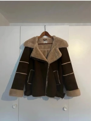 Brun shearling jacka från NA-KD - Snygg brun pälsjacka från NA-KD med beige fuskpäls på krage, ärmslut och insida. Jackan har dragkedja, två fickor framtill och dekorativa sömmar på ärmarna. Perfekt för kalla dagar och ger en cool look.