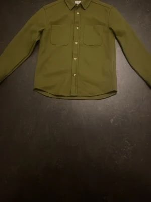 Oanvänd olivgrön overshirt med fickor - Stilren olivgrön overshirt med klassisk krage och två bröstfickor. Skjortan har knappar framtill och långa ärmar med knapp vid ärmslut. Materialet ser ut att vara tjockare och mjukt, perfekt för lager på lager. Enkel och clean design utan mönster.