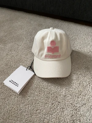 Vit keps från Isabel Marant - Snygg vit keps från Isabel Marant med broderad rosa logga och text framtill. Klassisk böjd skärm och justerbar passform bak. Tillverkad i ett slitstarkt tyg som ger en clean och trendig look. Perfekt accessoar för att lyfta din outfit.