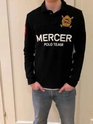 Svart Mercer Polo Team långärmad skjorta - Svart långärmad skjorta från Mercer Polo Team med vit text och emblem på bröstet samt röd siffra på ärmen. Klassisk krage och knappar framtill. Skjortan har en normal passform och är tillverkad i bomull, perfekt för en sportig och stilren look.