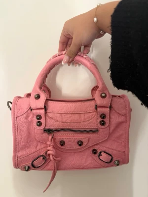 Rosa Balenciaga City handväska - Cool rosa Balenciaga City handväska i skinn med silverfärgade nitar och dragkedja framtill. Väskan har dubbla handtag, dekorativa spännen och klassiska detaljer som ger en edgy vibe. 