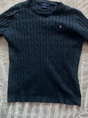 Mörkblå kabelstickad tröja Gant - Kabelstickad mörkblå tröja från Gant med rund halsringning och diskret logga broderad på bröstet. Tröjan har långa ärmar och ribbade muddar vid ärmslut och nederkant. Perfekt för en klassisk och stilren look.