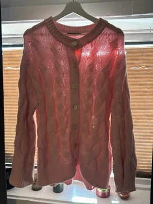 Rosa stickad kofta med knappar - Säljer en söt rosa stickad kofta med stora stickade flätor och ribbade muddar. Koftan har rund halsringning och knäpps framtill med vita knappar. Perfekt för dig som gillar mysiga och trendiga plagg i mjukt material.
