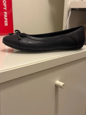 Svarta ballerinaskor från Geox - Stilrena svarta ballerinaskor från Geox i mjukt läder med klassisk rund tå och dekorativ rosett framtill. Skorna har en låg sula och diskreta sömmar för en enkel och tidlös look. Perfekta för dig som gillar minimalistisk stil.