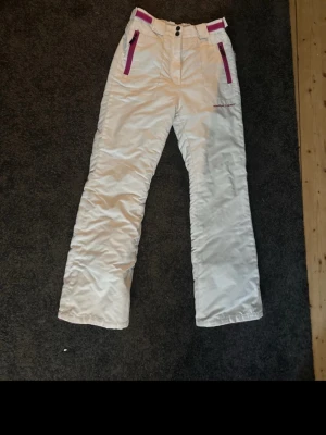Vita skidbyxor med rosa detaljer - Säljer ett par vita skidbyxor med raka ben och rosa dragkedjor vid fickorna. Byxorna har bälteshällor, knapp- och dragkedjestängning samt förstärkta sömmar. Perfekta för vintersport eller kalla dagar utomhus. En söm är sönder vid en av bälteshällorna. Säljer pga att de va för små tyvärr. Supersnygga💞