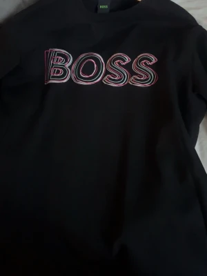 Svart sweatshirt från BOSS - Snygg svart sweatshirt från BOSS med stort, färgglatt BOSS-tryck på bröstet. Tröjan har en klassisk rund halsringning och ribbade muddar vid ärmslut och nederkant. Perfekt för en avslappnad och trendig look.