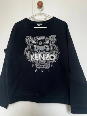 Svart sweatshirt med tiger från Kenzo - Svart sweatshirt från Kenzo med ikonisk broderad tiger och texten 'KENZO PARIS' på bröstet. Tröjan har rund halsringning och långa ärmar. Tillverkad i mjukt bomullsmaterial som är skönt att bära. Perfekt för dig som gillar streetwear och vill sticka ut.
