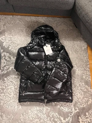 Svart glansig dunjacka från Moncler - Säljer en svart glansig dunjacka från Moncler med huva och dragkedja framtill. Jackan har två framfickor med dragkedja och Moncler-logga på ärmen. Perfekt för kalla vinterdagar och riktigt snygg streetstil.