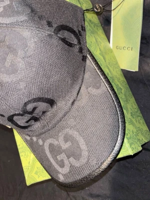 Svart Gucci keps med GG-monogram - Svart keps från Gucci med det ikoniska GG-monogrammet i ton-i-ton. Kepsen har böjd skärm och justerbar rem bak. Materialet är textil och kepsen levereras med original grön Gucci-box och tagg.