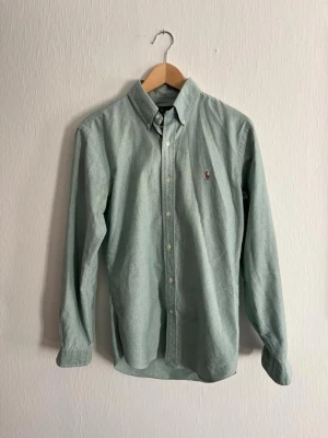 Mintgrön skjorta från Ralph Lauren - Säljer en mintgrön skjorta från Ralph Lauren i slim fit-modell. Skjortan har klassisk krage, knappar framtill och broderad logga på bröstet. Perfekt för dig som vill ha en stilren och snygg look. Materialet är bomull och den har långärmad design.